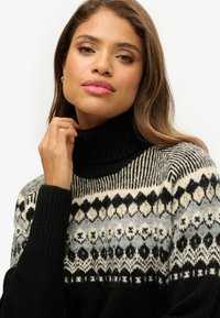Schwarzer Rollkragenpullover mit einem gemusterten Einsatz in Grautönen, Schwarz und Creme. Aus einem weichen, strukturierten Material gefertigt. Mit gerippten Bündchen.