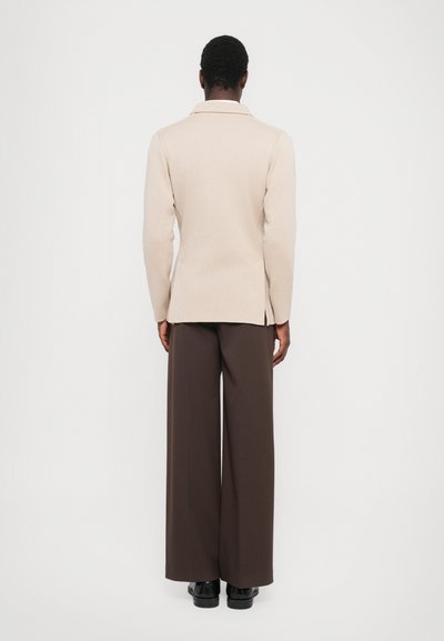 Beige Blazer mit Kragen, geradem Schnitt und seitlichen Schlitzen, kombiniert mit braunen weit geschnittenen Hosen und schwarzen Schuhen. Einfarbiger Hintergrund.