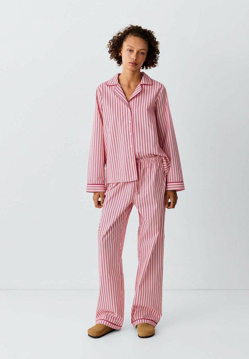 Femme debout portant un ensemble de pyjama rayé vertical rose et blanc avec boutons et pantalon large, assorti à des chaussons beige sur un fond uni.