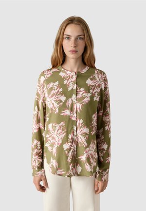 Junge Frau trägt eine olivgrüne Bluse mit langen Ärmeln und großen rosa-weißen Blumenmustern, kombiniert mit weißen Hosen, stehend vor neutralem Hintergrund.