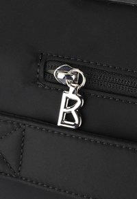 Sac en tissu noir avec une surface texturée, doté d'une tirette de fermeture éclair en argent avec une lettre "B" stylisée et des détails de couture contrastés.