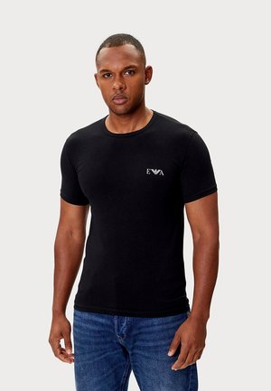 Zwart katoenen T-shirt met korte mouwen, een ronde hals en een klein wit logo op de linkerborst. Draagt blauwe spijkerbroek.