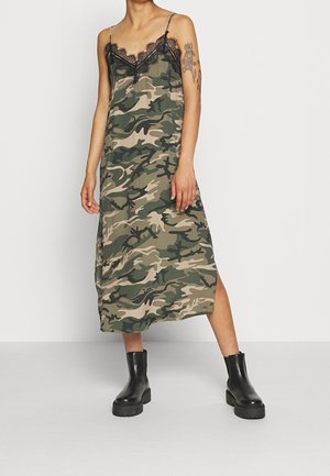 Nainen pukeutunut hihattomaan, maastokuvioiseen midimittaiseen mekkoon, jossa on mustaa pitsireunusta, ja mustiin nilkkureihin. Hän seisoo yksinkertaisen valkoisen taustan edessä.