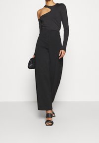 Top nero a maniche lunghe con taglio laterale, abbinato a pantaloni neri a vita alta e a gamba larga. Il modello tiene in mano una piccola borsa nera e indossa sandali neri.