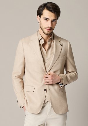 Młody mężczyzna o ciemnych włosach i jasnym zarostem, ubrany w beżową marynarkę, kamizelkę i koszulę, zapinający guzik marynarki na neutralnym tle.