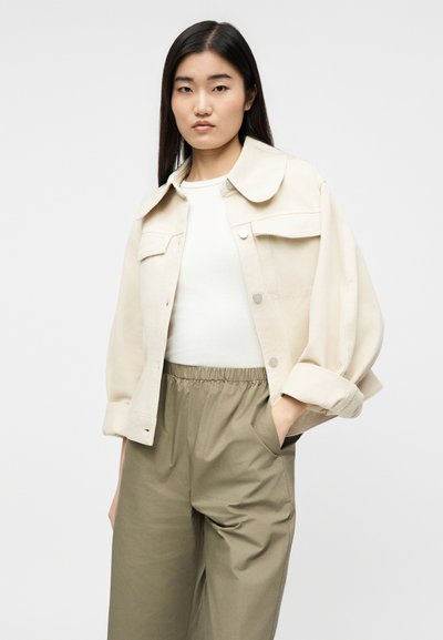 Γυναίκα με μακριά σκούρα μαλλιά, που φοράει ένα κρεμ κοντό μπουφάν, λευκό τοπ και φαρδιά χακί παντελόνια με τα χέρια στις τσέπες.