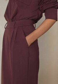 Bourgognerøde bukser med glat tekstur, høj talje, sidelommer og markante læg. Har en matchende bæltedetalje.