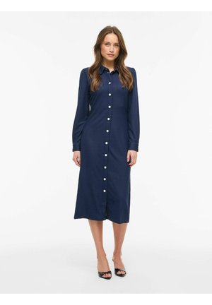 Kvinde stående iført en navyblå, langærmet, knap-ned midi-kjole med sorte åbne hæle på hvid baggrund.