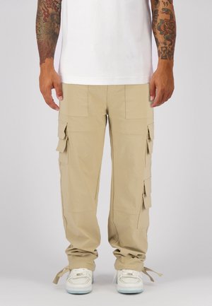 Beige cargobroek met meerdere zakken, recht model, verstelbare manchetten en een lichte stof, gecombineerd met witte sneakers.