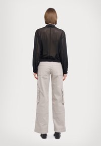 Blusa negra transparente de manga larga con puños fruncidos, combinada con pantalones cargo de color gris claro que cuentan con bolsillos laterales y un ajuste relajado.