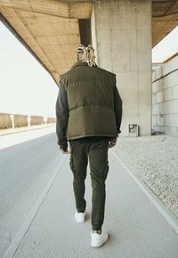 Gilet matelassé vert olive sur une chemise noire à manches longues, associé à un pantalon cargo olive et des baskets blanches, vu de dos.