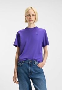Purple cotton t-shirt with a crew neckline and short sleeves. Features a small emblem on the chest, paired with blue denim jeans.
