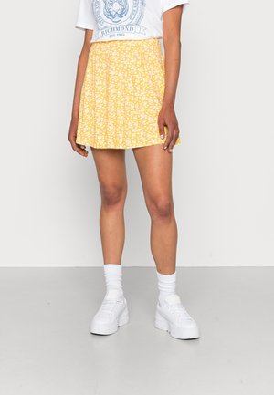 Persoon die een gele minirok met bloemenprint draagt, een wit T-shirt met opdruk, witte plateau sneakers en witte crewkousen, tegen een effen achtergrond.