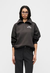 Σκούρο γκρι φούτερ με φερμουάρ και ψηλό γιακά, με το λογότυπο «NEW BALANCE». Συνδυάζεται με μαύρο παντελόνι, που έχει άνετη εφαρμογή και τσέπες.