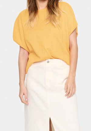 Femme portant un haut jaune à manches courtes ample, rentré dans une jupe blanche taille haute avec une fente à l'avant et une fermeture par boutons.