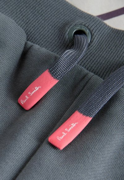 Close-up af grå stofsnore med lyserøde ender mærket "Paul Smith" på et mørkegråt strikket tøjstykke.