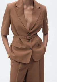 Blazer marrone su misura con maniche corte, revers a punta, due tasche frontali e una cintura coordinata. Tessuto liscio con vestibilità strutturata.