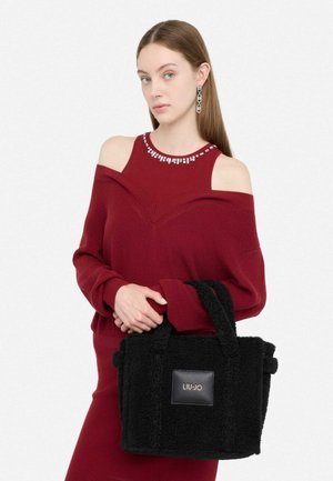 Borsa a tracolla nera in peluche con manici corti e forma rettangolare, dotata di una patch in pelle con logo. Modello indossa un maglione bordeaux off-shoulder.