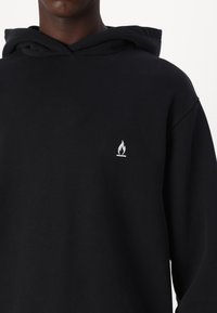 Haut à capuche noir avec une poche kangourou sur le devant, capuche à cordon de serrage et un petit logo de flamme blanche sur la poitrine gauche. Tissu doux et texturé.