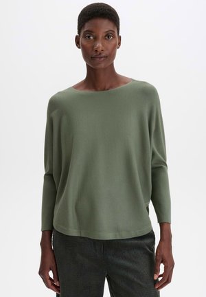 Donna che indossa un maglione verde oliva a maniche lunghe e largo e pantaloni scuri con trama, in piedi davanti a uno sfondo bianco semplice.