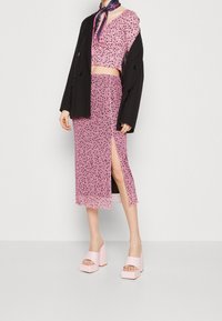 Žena v růžovém květinovém crop topu s odpovídající midi sukní se bočním rozparkem, přehozeným černým oversized blejzrem, fialovým šálem na krku a růžovými platformovými podpatky.