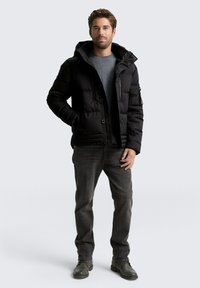 Černá péřová bunda s kapucí, se zipovou kapsou, háčkovými uzávěry a texturovanou tkaninou. V kombinaci se šedými džíny a robustními botami.