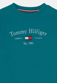 Teal bomuldstrøje med broderet tekst "Tommy Hilfiger" og logo, med rund hals og ribbet krave. Klassisk pasform.