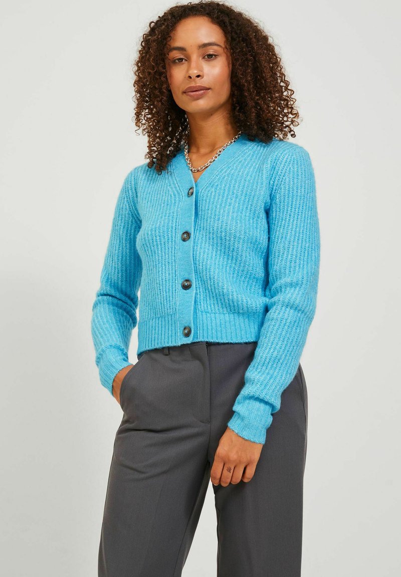 Cardigan a coste di colore azzurro chiaro con scollo a V, chiusura frontale con bottoni e maniche lunghe, abbinato a pantaloni grigi. Materiale in maglia testurizzata.