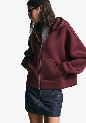 Bordo zip-up kapucar s sproščenim krojem, z velikima sprednjima žepoma in kapuco na vrvico, v kombinaciji z denim mini krilom.