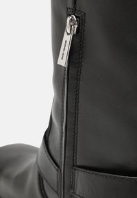 Botte en cuir noir avec une fermeture éclair sur le côté dotée d'un tirette en argent, des détails de couture et une texture lisse.
