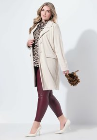 Donna che cammina con un lungo cappotto beige, un top con stampa leopardata, leggings in pelle bordeaux e tacchi bianchi, mentre tiene in mano una piccola borsa frangiata marrone.