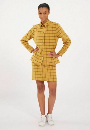 Femme debout, les mains sur les hanches, portant une chemise à carreaux jaune moutarde boutonnée et une jupe assortie, associées à des baskets blanches.