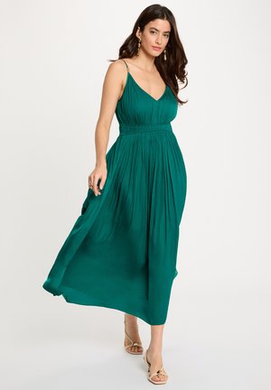 Støvet blågrøn ærmeløs kjole med V-hals og taljeret snit. Flydende stof, knælang, med plissé og subtile guldaccents på stropperne.