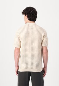 Polo din tricot cu mânecă scurtă, într-o nuanță deschisă de bej, având un model texturat cu detalii din țesătură deschisă și un guler și un tiv cu nervuri.