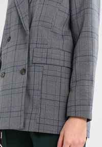 Harmaa ruudullinen bleiseri, jossa on tiklihahmokuvio, kaksi ruskeaa nappia ja yksi tasku. Kankaan pinta näyttää sileältä ja istuu räätälöidysti.