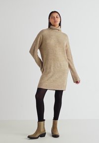 Beige kötött garbóruha hosszú ujjakkal, laza fazonnal és bordázott textúrával, fekete harisnyával és bézs bokacsizmával párosítva.