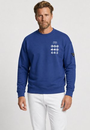 Man met baard die een blauwe trui draagt met doorgestreepte voetbalformaties en tekst "TOTAL FOOTBALL" op de borst, witte broek, neutrale achtergrond.