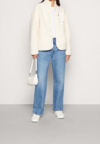 Kość słoniowa kurtka z polaru z zapinaną na suwak kieszenią na piersi, zestawiona z niebieskimi jeansami o prostych nogawkach i białą mini torebką. Całość dopełniają białe sportowe buty.