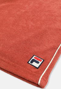 Tissu rouge texturé avec un passepoil blanc sur le bord et un petit patch logo carré en bleu marine, blanc et rouge près du coin.