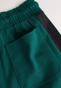 Primer plano de pantalones verdes con cintura elástica, franja lateral negra y un gran bolsillo cuadrado trasero.