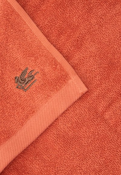 Serviette orange corail douce avec un coin plié montrant un petit logo d'oiseau gris brodé près du bord.