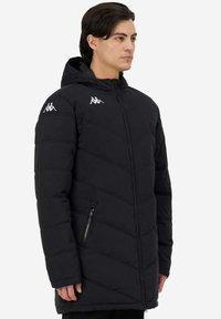 Giacca nera imbottita con cappuccio, caratterizzata da cuciture diagonali, tasca laterale con zip e accenti bianchi del logo sulle spalle.