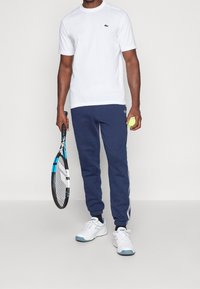 Man i vit t-shirt, marinblå joggers och vita sneakers som håller en tennisklubba i ena handen och en tennisboll i den andra.