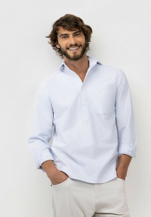 Joven con cabello castaño y barba sonriendo, lleva una camisa a rayas azul claro con las mangas remangadas y pantalones de color claro, manos en los bolsillos.