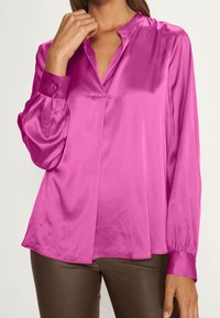 Blouse en satin fuchsia avec col montant, manches longues bouffantes et poignets boutonnés, dotée d'un pli frontal pour une coupe décontractée.