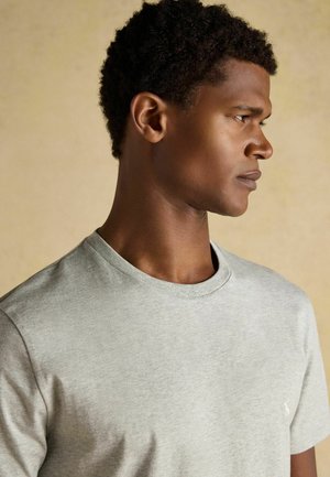 Nuori mies, jolla on lyhyet kiharat hiukset, pukeutuneena vaaleanharmaaseen pyöreäkaulaiseen t-paitaan, katsoo oikealle yksiväristä beigeä taustaa vasten.