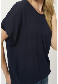 Femme portant un chemisier ample bleu marine à manches courtes et un jean bleu, avec de longs cheveux blonds partiellement visibles.