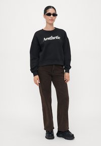Felpa nera con la scritta "Aesthetic", fit ampio, polsini a coste, abbinata a jeans marroni a gamba larga e sneakers nere chunky.
