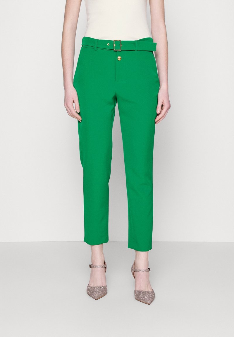 Pantalon vert sur mesure avec une taille ceinturée, des détails en métal doré, et une coupe courte, associé à des escarpins pailletés à bouts pointus.