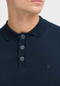Polo shirt bleu marine foncé avec un col à côtes, patte de boutonnage à quatre boutons de couleur marine et un petit emblème brodé sur la poitrine.